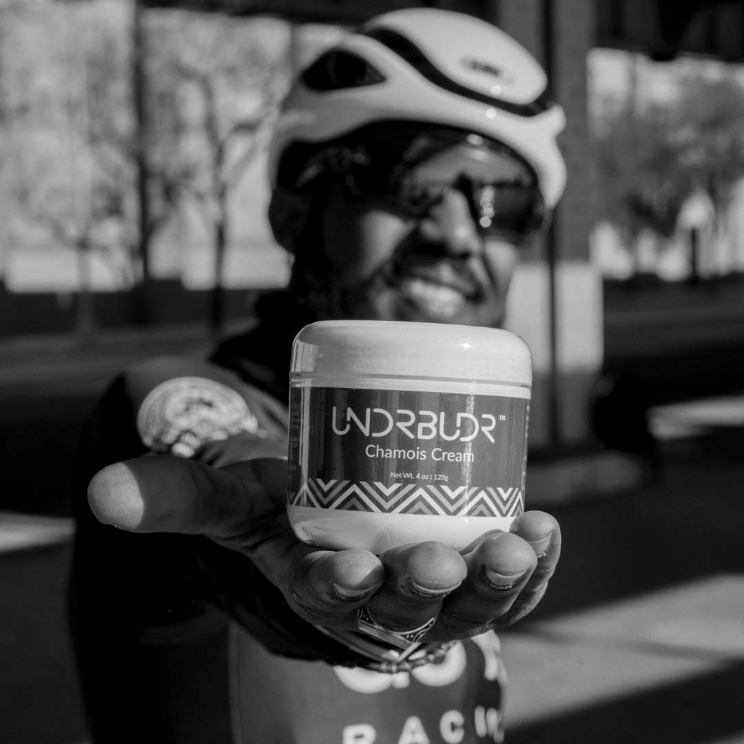 Male cyclist holding a tub of UNDRBUDR chamois cream up to the camera.