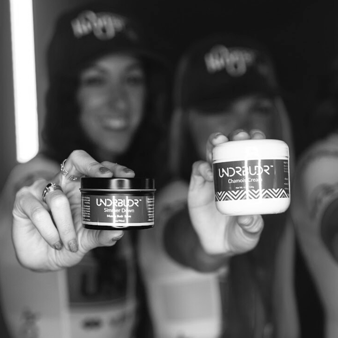 Two women cyclists holding UNDRBUDR chamois cream and muscle rub up to the camera.